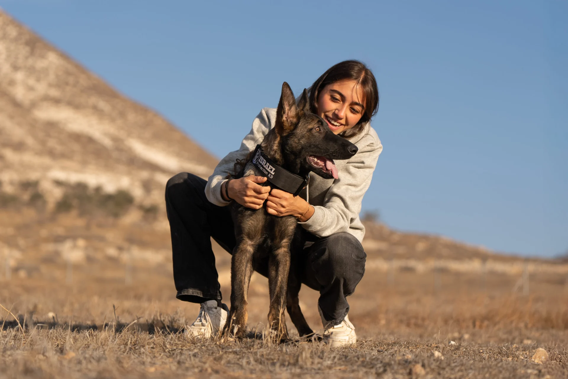 Woman kneeling beside a Belgian Malinois in training, showcasing a bond between handler and protection dog in a natural outdoor setting.