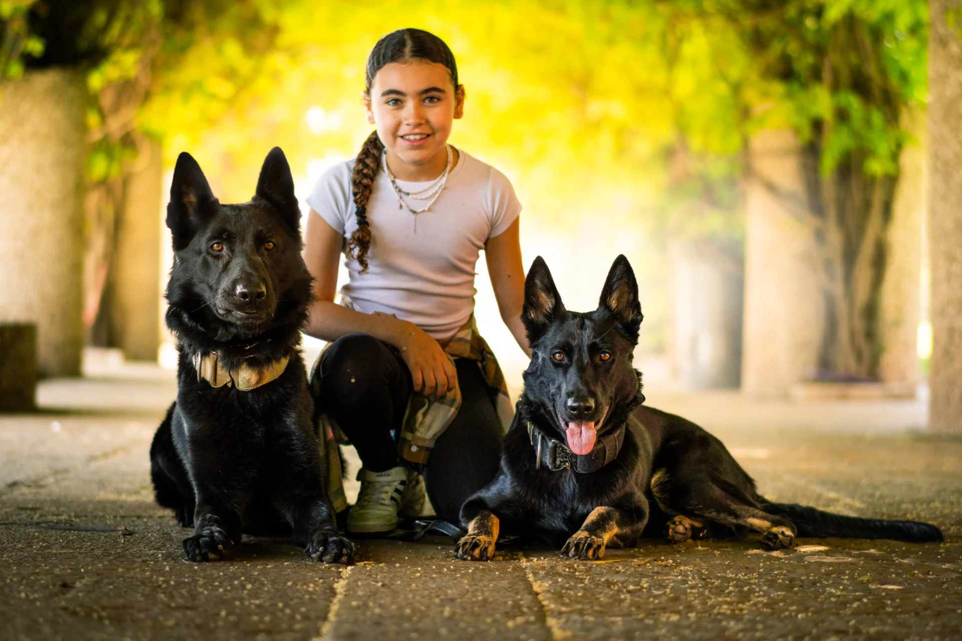 Girl sitting with two trained black protection dogs in a natural outdoor setting, showcasing the bond and integration between the handler and K9s as part of their immersive training experience.