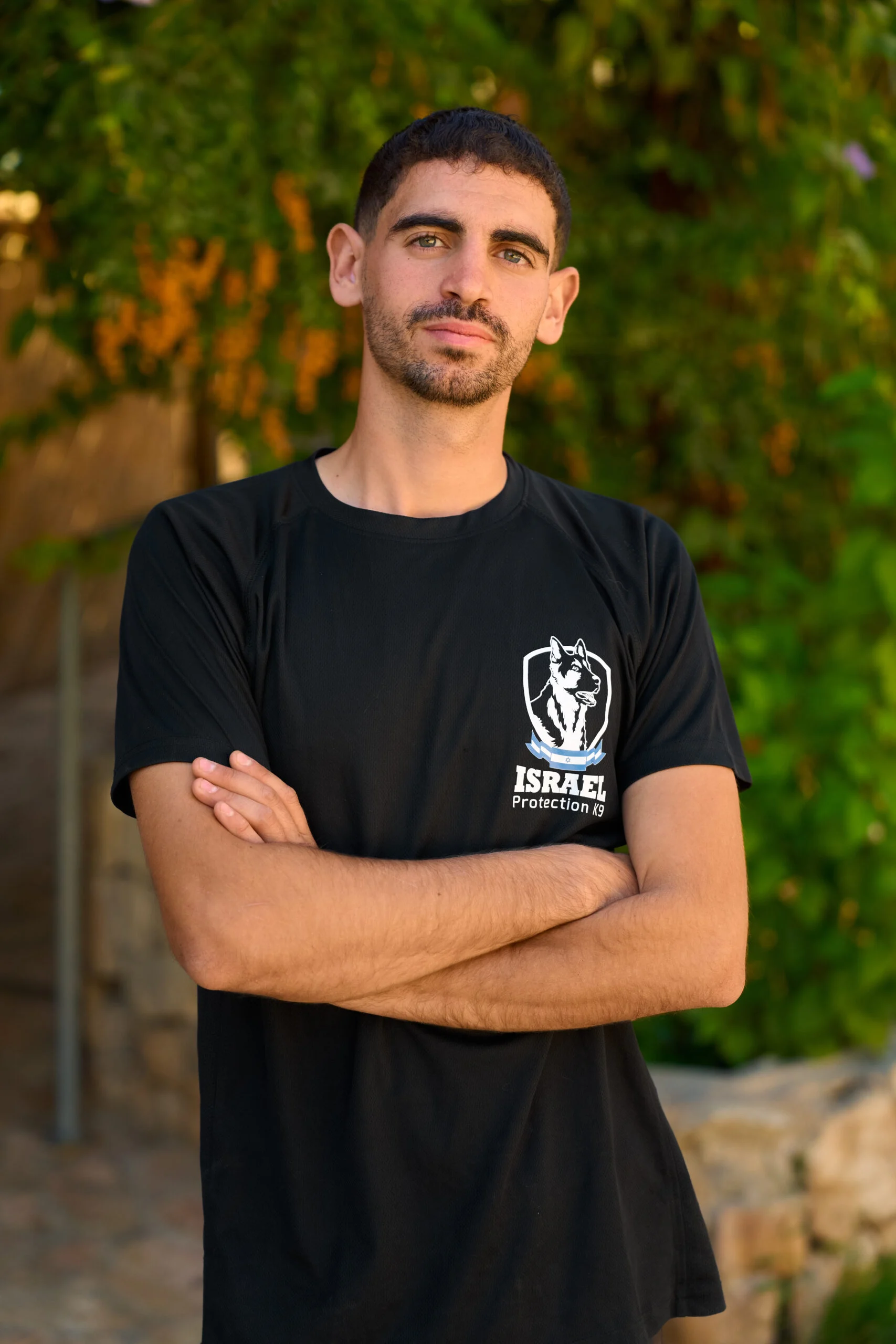 Yosef Biton, Israel Protection K9 trainer, standing with crossed arms against a green background.